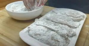 Hand coating chicken fillets in flour on a white plate for frying on a wooden countertop.